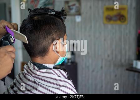 Teenager in einer Gesichtsschutzmaske bekommt einen Haarschnitt von einem Friseurladen. Modisch längliche Haarschnitte für Jungen. Schönheitssalon in Quarantäne coro Stockfoto