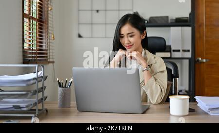 Die charmante und schöne, tausendjährige asiatische Geschäftsfrau oder Marketingmanagerin sitzt lächelnd an ihrem Schreibtisch und blickt zufrieden auf ihren Laptop-Bildschirm Stockfoto