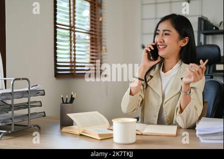 Die charmante und attraktive asiatische Geschäftsfrau unterhält sich gerne mit ihrem Kollegen am Telefon, während sie an ihrem Schreibtisch sitzt. Stockfoto