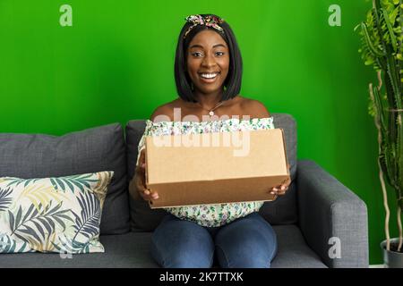 Afrikanische junge Frau sitzt auf der Couch zu Hause Auspacken Karton mit Online-Kauf Stockfoto