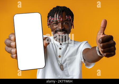 Aufgeregt junger afrikanischer Mann hält Smartphone mit weißem leeren Bildschirm in der Hand auf gelbem Studiohintergrund Stockfoto