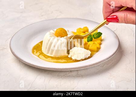 Frauenhand mit Löffel bricht ein Stück hausgemachte Panna Cotta mit natürlicher Vanilleschote, Mangosirup und Minze, selektiver Fokus Stockfoto