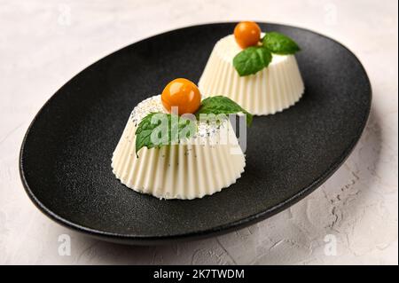 Zwei Panna Cotta mit natürlicher Vanilleschote, Physalis oder Winterkirsche und Minze auf schwarzem ovalem Teller, Nahaufnahme Stockfoto