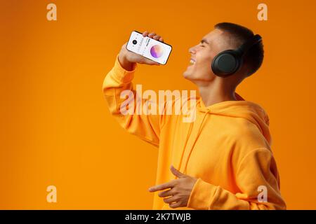 Junger gutaussehender Mann mit Smartphone als Mikrofon vor gelbem Hintergrund Stockfoto