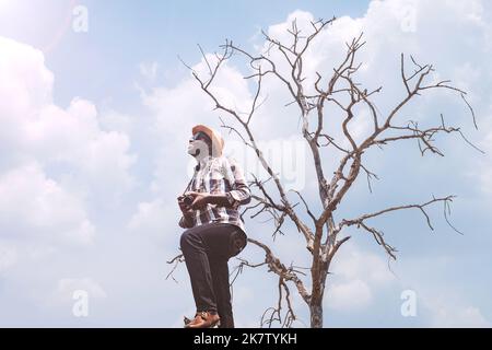 Afrikanischer Fotograf, der eine Kamera verwendet, während er eine Savanne mit dem trockenen Baum und dem Himmel im Hintergrund fotografiert Stockfoto