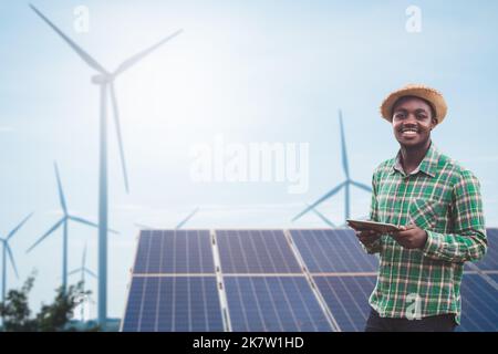 Afrikanischer Landwirt steht und hält digitale Tablette auf Maisfarm mit Solarzelle und Windturbine im Hintergrund.Konzept der Ökostrom Nachhaltigkeit r Stockfoto