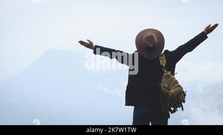 Ein männlicher Reisender in einem Rucksack steht oben auf dem Berg mit den Händen nach oben und zeigt an einem nebligen und regnerischen Tag das Zeichen der Freiheit. Abenteuer und Stockfoto