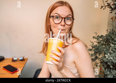 Portrait eines hübschen jungen Mädchens, das im Hintergrund eines Stadtcafés aus gelbem Plastiksodabecher trinkt Stockfoto