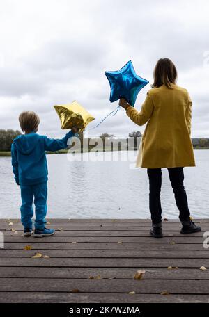 Junge und Frauen in gelb-blauen Kleidern mit Ballons stehen mit Rücken in der Nähe des Sees. Flüchtlinge, Einheit, Unterstützung, Nostalgie, Traurigkeit. Die Ukrainer sind tu Stockfoto