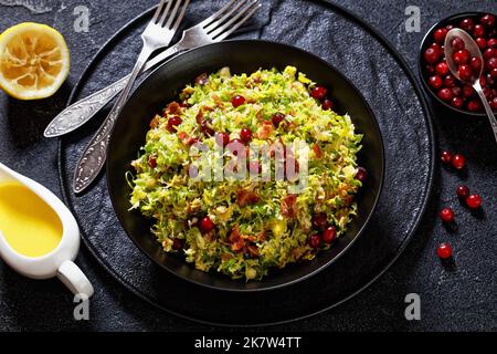 brüsseler Sprossen schlagen mit Mandeln, knusprig gebratenem Speck und Preiselbeeren in einer schwarzen Schüssel auf einem Betontisch mit Zutaten Stockfoto