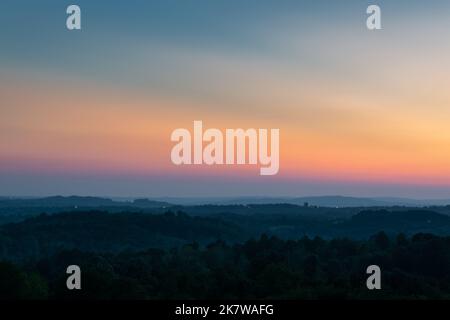 Hügelschichten, die bei Dämmerung durch Dunst und strahlendes Leuchten am Himmel über dem Horizont getrennt sind, hügelige ländliche Landschaft mit dem letzten Licht am klaren Himmel Stockfoto