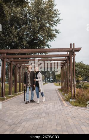 In voller Länge ein glückliches junges Paar in herbstlichen Mänteln, das im Park unter mehreren Bögen läuft, Stockbild Stockfoto