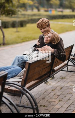 Fröhlicher junger Mann umarmt positive Frau und sitzt auf Holzbank im Park, Bild Stock Stockfoto