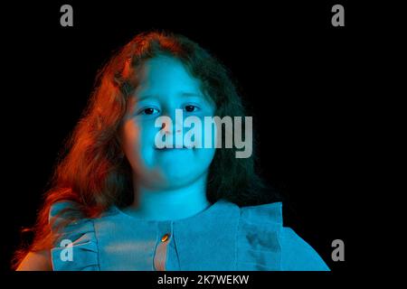 Charmante rothaarige kleine Mädchen trägt festliches Kleid Blick auf Kamera isoliert über dunklen Hintergrund in Neonlicht. Kinder Emotionen, Gesichtsausdrücke Stockfoto