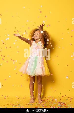 Lustige kleine Mädchen für Party in Pailletten und Tutu weht Konfetti gekleidet. Isoliert auf gelbem Hintergrund. Stockfoto