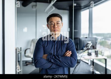 Porträt eines ernsthaften asiatischen Entwicklers, eines Geschäftsmannes mit gekreuzten Armen, der konzentriert auf die Kamera schaut, eines Mannes in Brille und Hemd, der im modernen Bürogebäude arbeitet. Stockfoto
