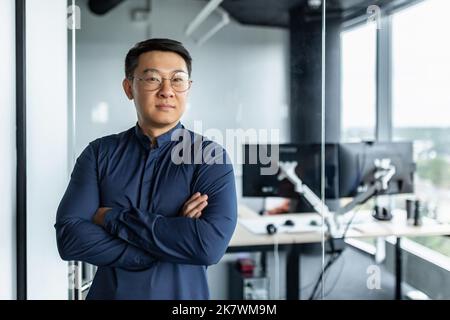 Porträt eines Geschäftsmannes mit gekreuzten Armen, eines asiatischen Mannes, der lächelt und die Kamera anschaut, eines Mannes, der im modernen Bürogebäude arbeitet. Stockfoto