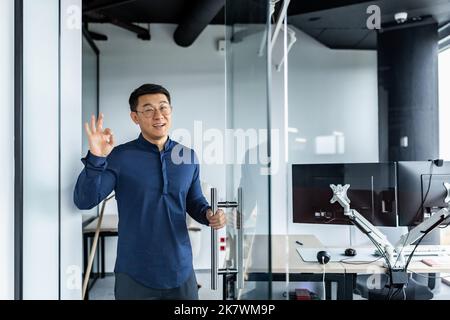 Ein asiatischer Geschäftsmann schaut auf die Kamera und lächelt, der Mann zeigt das Schild ok alles ist in Ordnung, der Chef lädt ihn in sein Büro ein. Stockfoto