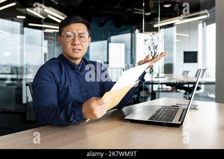 Verärgerter asiatischer Geschäftsmann, der die Kamera ansah und einen Brief mit schlechten Nachrichten zeigte, einen Mann, der im modernen Bürogebäude arbeitete und einen Laptop benutzte. Stockfoto