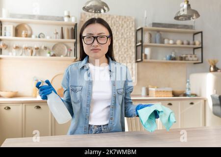 Porträt einer unzufriedenen jungen asiatischen Frau, Hausfrau in einer Brille, die auf die Kamera schaut, die Waschmittel und Schwamm hält. Stockfoto