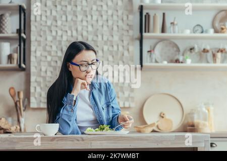 Unzufrieden junge asiatische Frau in Brille und Jeanshemd sitzen in der Küche zu Hause am Tisch. Er versucht einen frischen Salat mit einer Gabel, sieht ihn an, müde von einer Diät zu sein. Stockfoto