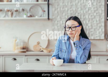 Kaffeepause. Junge müde asiatische Frau Freiberufler. Sitzen zu Hause am Küchentisch mit einer Tasse Kaffee, ausruhen, nachdenklich. Stockfoto