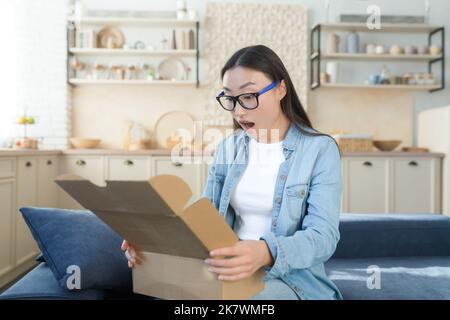 Schockiert und fröhlich Frau zu Hause erhielt einen freudigen Kauf eines Geschenks, öffnet die Box und untersucht das Geschenk mit Befriedigung, sitzt auf dem Küchensofa. Stockfoto