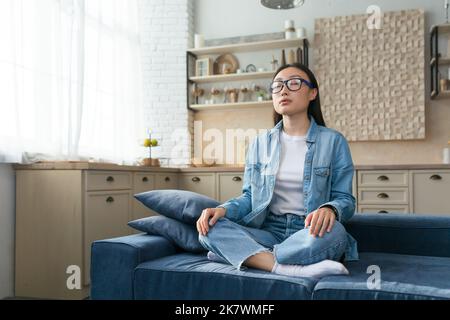 Yoga zu Hause. Eine junge Asiatin in Gläsern sitzt ruhig und entspannt zu Hause auf dem Sofa in der Lotusposition, schloss ihre Augen und meditierte. Stockfoto