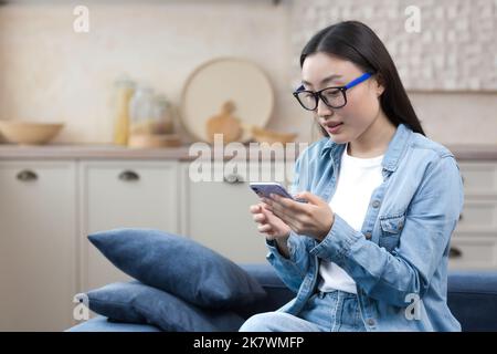 Junge schöne Frau zu Hause sitzt in der Küche lächelnd und glücklich, asiatische Frau mit Smartphone Nachrichten tippen und Nachrichten online lesen. Stockfoto