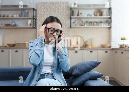 Die junge kranke Frau zu Hause hat starke Kopfschmerzen, die asiatische Frau sitzt auf dem Sofa in der Küche und spricht am Telefon, ruft den Arzt um Hilfe und konsultiert. Stockfoto