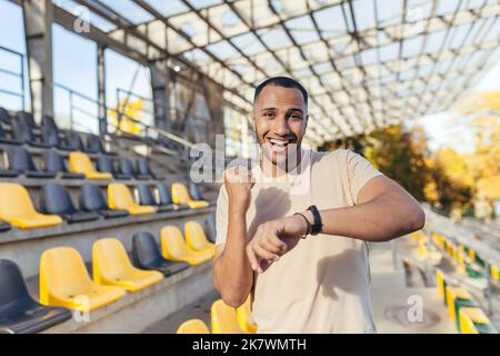 Glücklicher junger Sportler, der die Kamera anschaut und glücklich lächelt mit einem guten Fitnessergebnis, Mann, der die Zeit auf einer intelligenten Uhr des Fitnessarmbandes überprüft, tagsüber im Sportstadion läuft. Stockfoto