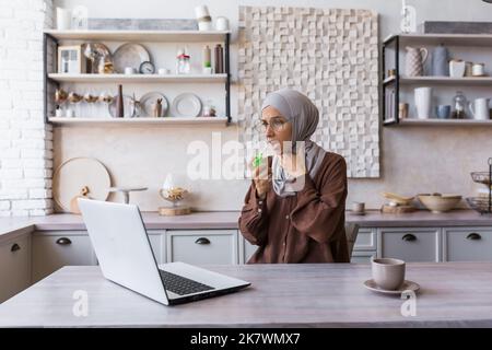 Kranke muslimische Frau in Hijab zu Hause, Frau hat starke Halsschmerzen Halsschmerzen, verwendet Schmerzspray zu Hause in der Küche sitzend. Stockfoto
