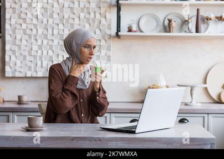 Kranke muslimische Frau in Hijab zu Hause, Frau hat starke Halsschmerzen Halsschmerzen, verwendet Schmerzspray zu Hause in der Küche sitzend. Stockfoto