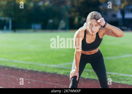 Müde Sportlerin, die sich nach dem Joggen an einem sonnigen Tag im Stadion ausruhte, blonde Sportkleidung, die eine Verschnaufpause einnahm. Stockfoto