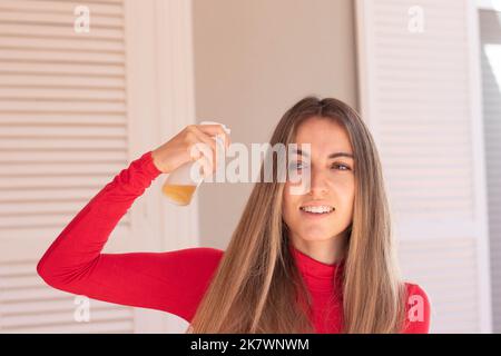 Vorderansicht einer jungen schönen Frau, die ätherische Öle aus einer Glasflasche in ihr gesundes langes Haar sprüht und wegschaut Stockfoto