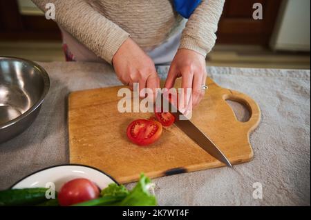 Blick von oben auf eine Hausfrau, die Tomaten auf einem Holzbrett hackt und in der heimischen Küche gesunden, rohen veganen Salat zubereitet Stockfoto