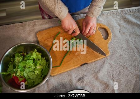 Draufsicht : Hände mit Küchenmesser, hacken junge grüne Zwiebel auf einem Holzbrett, Vorbereitung gesunder Gemüsesalat. Stockfoto