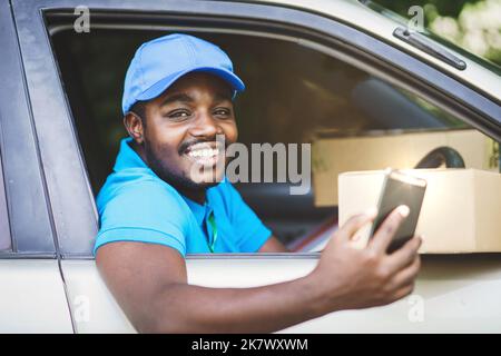 Afrikanischer Kurier der Lieferfirma mit Smartphone und hält ein Paket im Auto mit Lächeln und glücklich.Konzept der Business Delivery Service Stockfoto