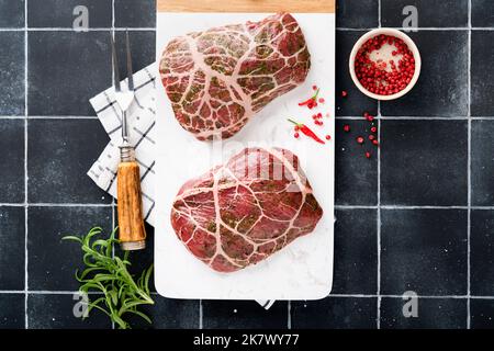 Steaks Raw. Rohes Cowboy-Steak, marmoriertes Rindfleisch mit Gewürzen Rosmarin und Pfeffer auf weißem Marmorschiefer und grauem Tischhintergrund. Draufsicht. Modell. Stockfoto