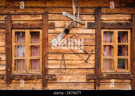 Eine alte Holzwand eines Hauses mit Fenstern im Dorf und Werkzeugen Stockfoto