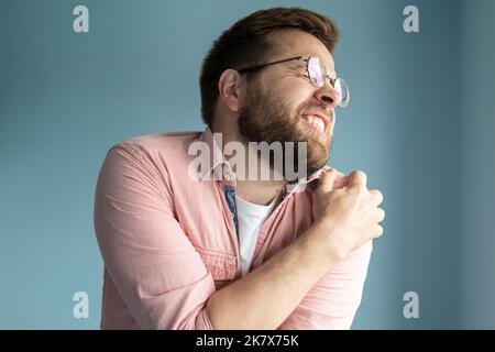 Der junge Mann hat starke Schmerzen in der Schulter und massiert mit Leiden im Gesicht die Wunde Stelle. Medizinisches Konzept. Blauer Hintergrund. Stockfoto