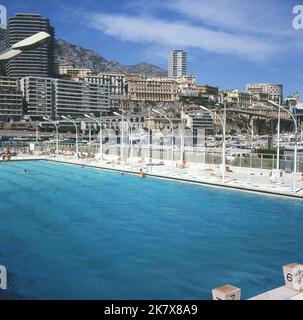 1964, historisch, ein Blick aus dieser Zeit auf den Staat Nautique Rainier III, ein Freibad in der Mitte des Port Hercules in Monaco. Der Meerwasserpool in olympischer Größe wurde im August 1961 offiziell eröffnet. Die luxuriösen Apartmentblöcke mit Blick auf den Hafen sind in der Ferne zu sehen. Stockfoto