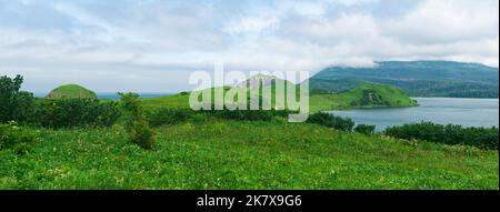 Natürliche Landschaft der Insel Kunashir mit grasbewachsenen Hügeln, vulkanischen Felsen, Vulkan in den Wolken und einem Tal mit einer Lagune Stockfoto