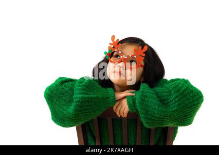 Cute asiatische Kind Mädchen in grünen Pullover und Maskerade weihnachtsgläser mit Hirsch Hörner. Portrait schöne Thai-Mädchen suchen nach oben auf die Seite trägt ein Stockfoto