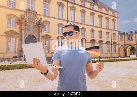 Glücklicher Student Mann in der Nähe des Universitätscampus mit einer deutschen Flagge und das Erlernen der Sprache über das Internet auf einem Laptop Stockfoto