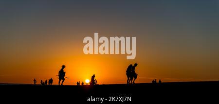 Ein Panoramablick auf Silhouetten einer Gruppe von Menschen Spaziergang entlang der Küste bei einem wunderschönen Sonnenuntergang Stockfoto