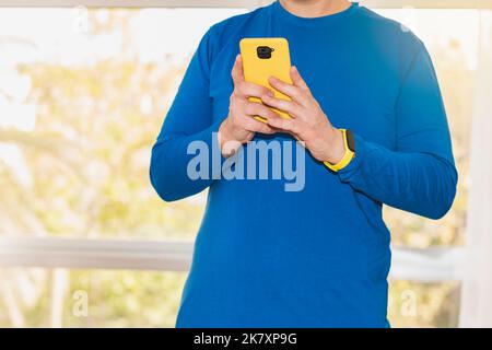 Ein unbekannter kaukasischer Mann, der in einem blauen Langarm-T-Shirt gekleidet ist, verwendet ein Smartphone mit einem gelben Gehäuse. Er trägt ein Aktivitäts-Armband. Stockfoto