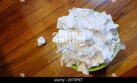 Blick von oben auf einen kleinen Mülleimer voll mit gebrauchten Papiertaschentüchern, auf Holzboden. Stockfoto