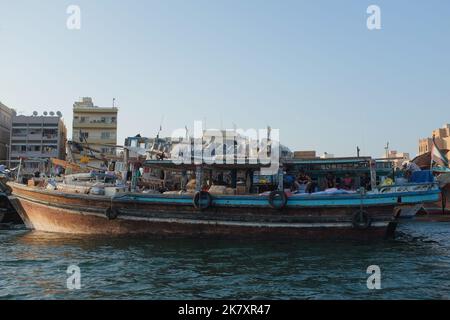 Altes und traditionelles Frachtschiff, genannt Dhow, vertäut am Dubai Creek, Gebäude von Deira dahinter, in Dubai, VAE. Männer hängen an der Seite des Bootes herum. Stockfoto