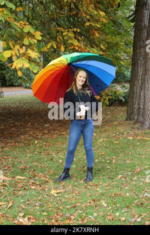 Attraktive Frauen aus Kaukasien mittleren Alters in Belleisle Park, Ayr, Ayrshire, Schottland, Großbritannien Genießen Sie die Herbstfarben in Jeans und schwarzem Wolltop und tragen einen bunten Regenbogen-Regenschirm mit Brolly Stockfoto